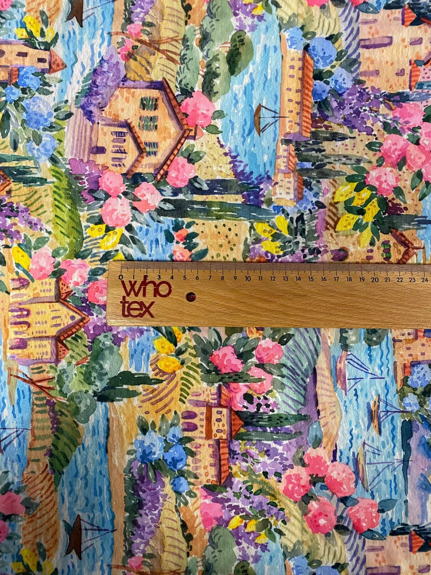 Watercolor coastal village print fabric with colorful houses, flowers, and water details, shown with wooden ruler for scale