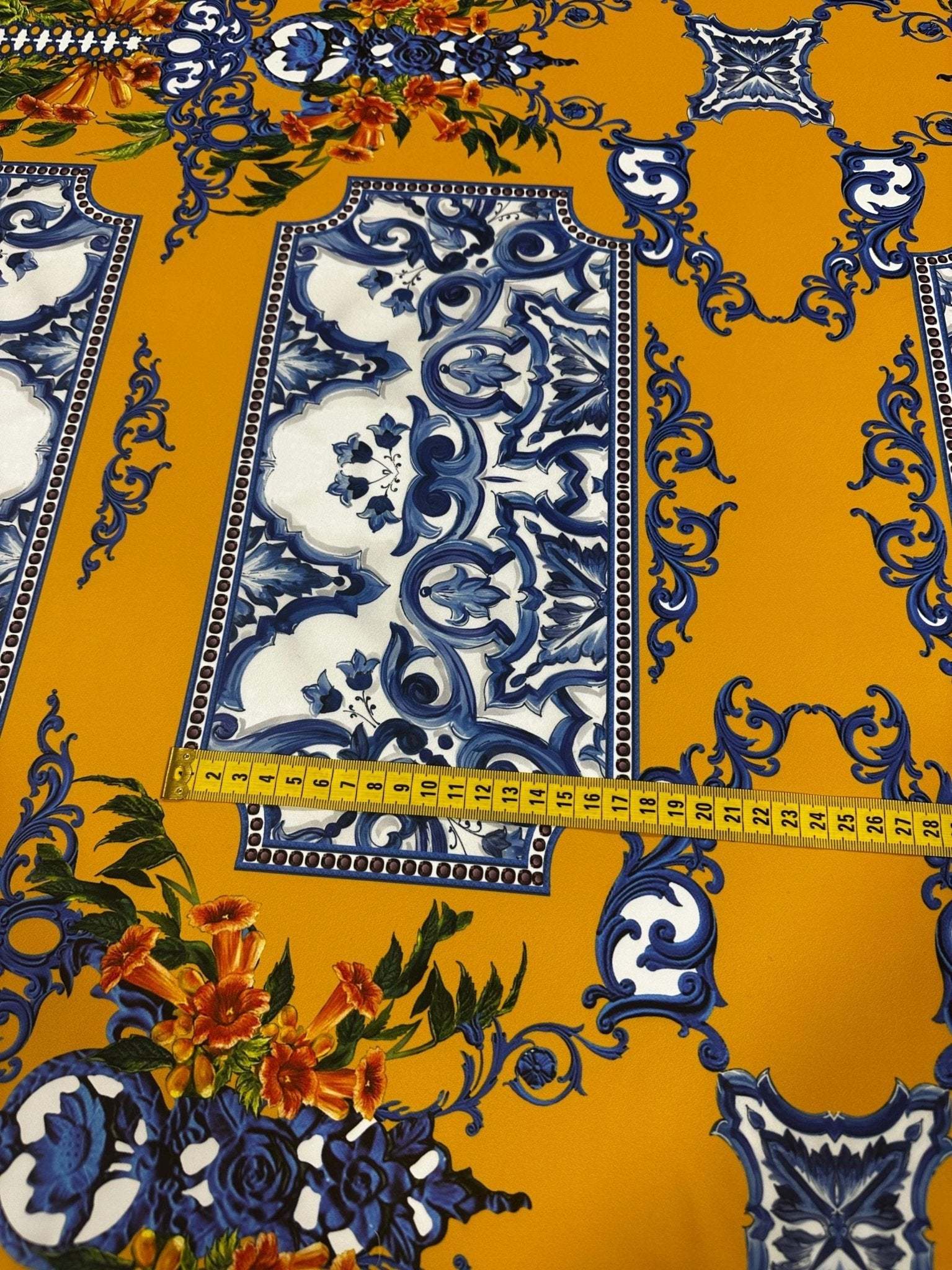 Elegant mustard yellow Baroque floral fabric with intricate blue and white patterns and colorful floral accents, featuring a measuring tape showing fabric width.