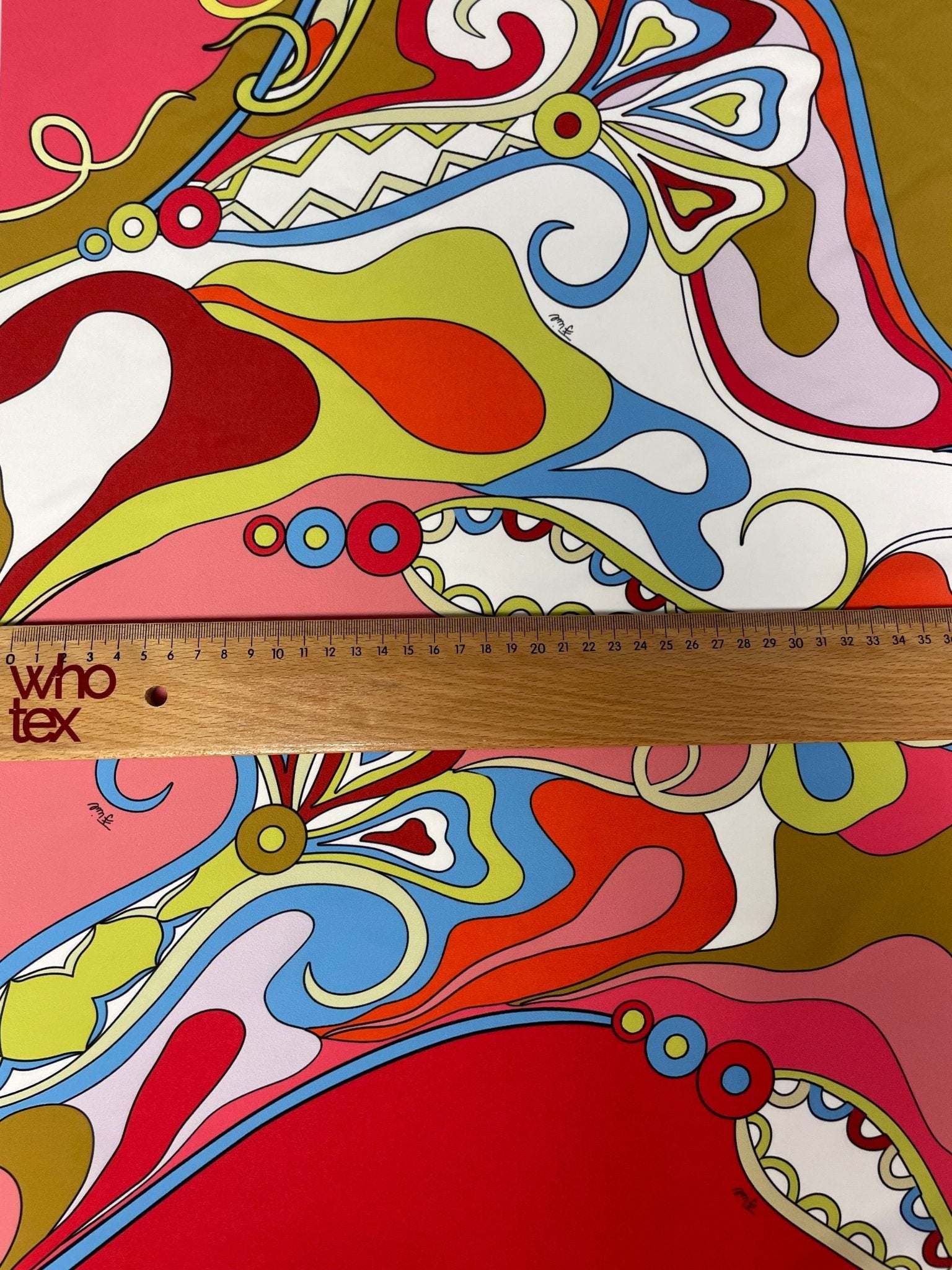 colorful abstract swirl pattern fabric with vibrant red, blue, yellow, and green swirls and shapes, shown with a wooden ruler for scale