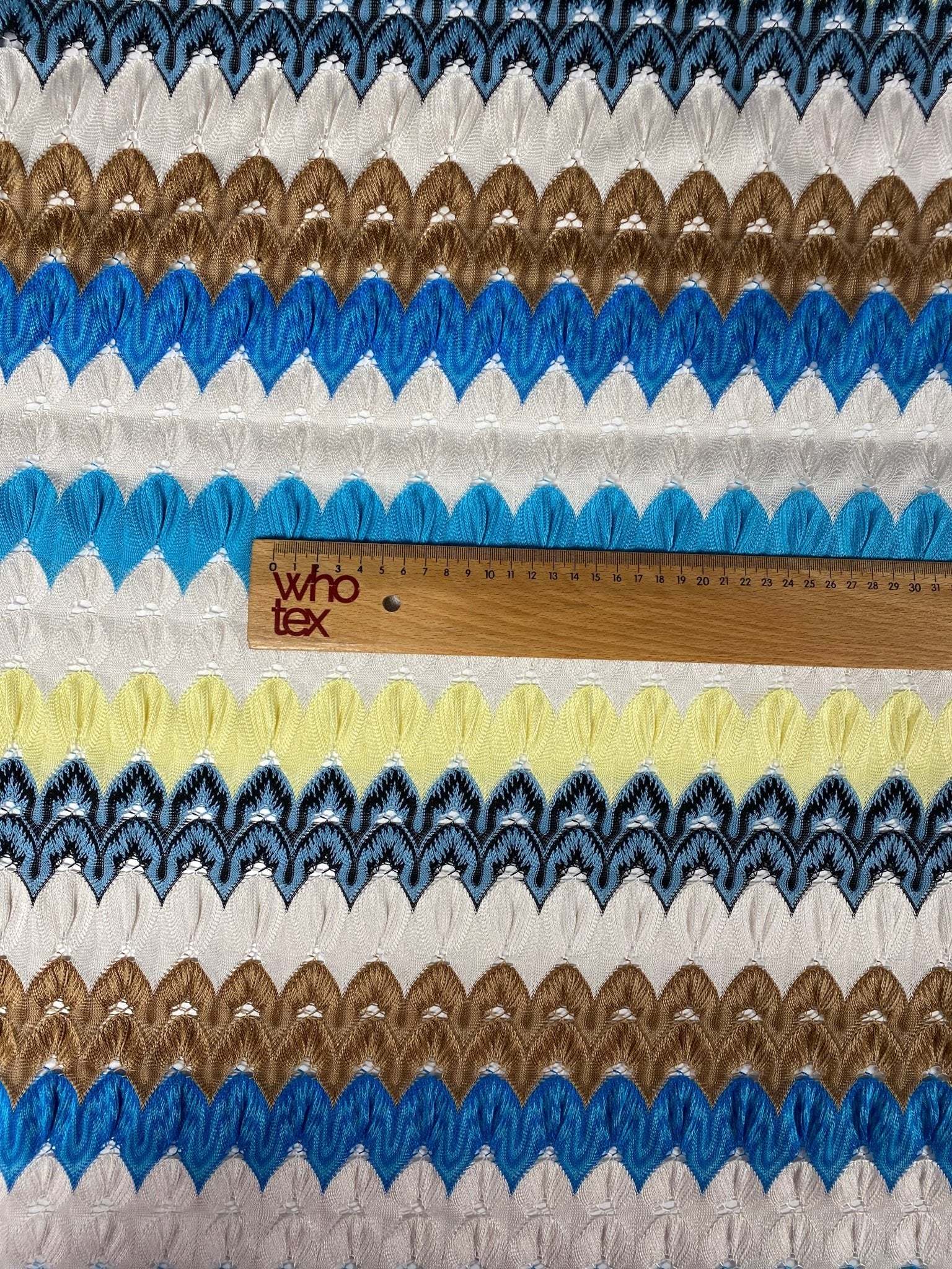 Close-up of colorful chevron embroidered fabric with zigzag knit pattern in blue, brown, yellow, white, and black tones with a wooden ruler for scale