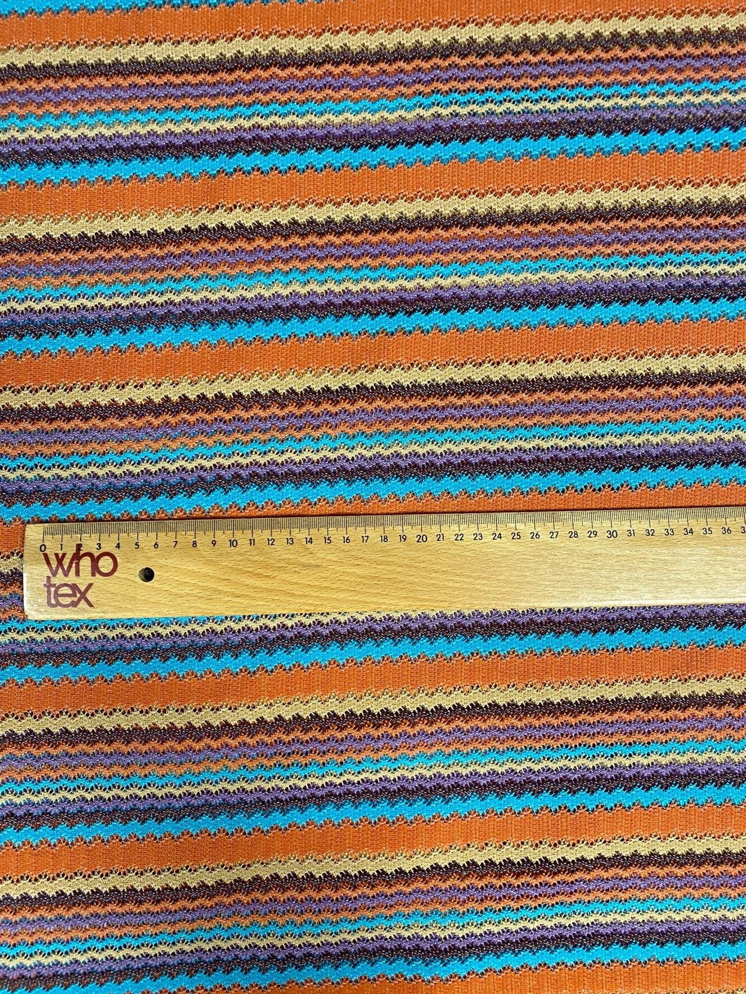 Colorful zigzag stripe knit fabric in green, orange, blue, and brown tones with a wooden ruler for scale