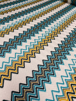 Close-up of geometric zigzag embroidered fabric in green, teal, mustard, and white tones with intricate knit texture