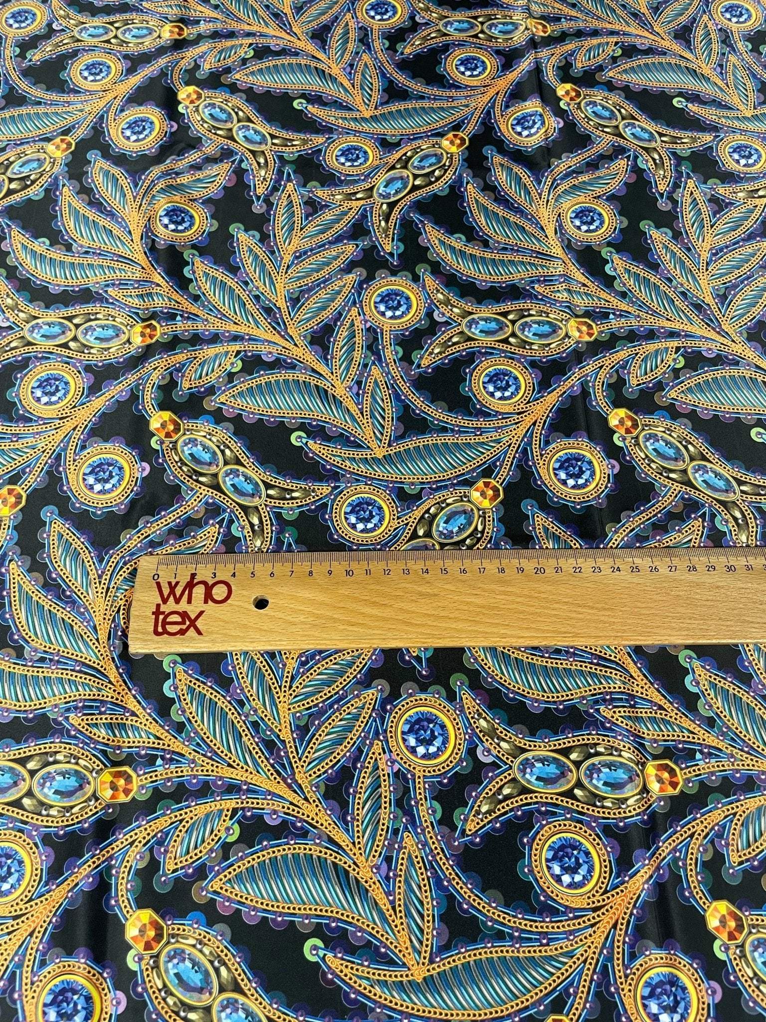 luxury peacock feather fabric with jewel accents and intricate blue and gold patterns on dark background with ruler for scale