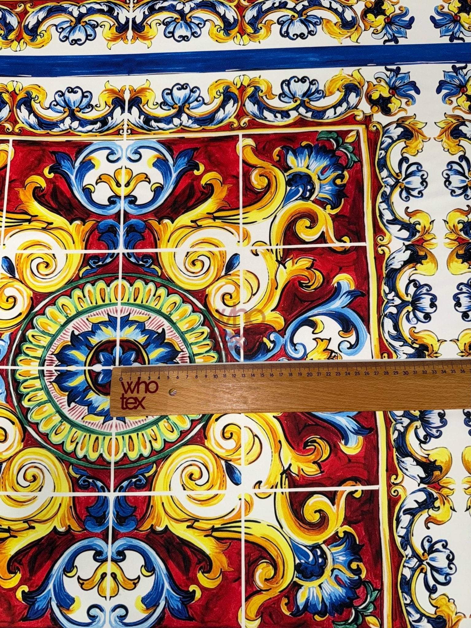 Close-up of Majolica Pattern Luxury Fabric featuring vibrant red, yellow, blue, and white intricate designs with a ruler for scale.