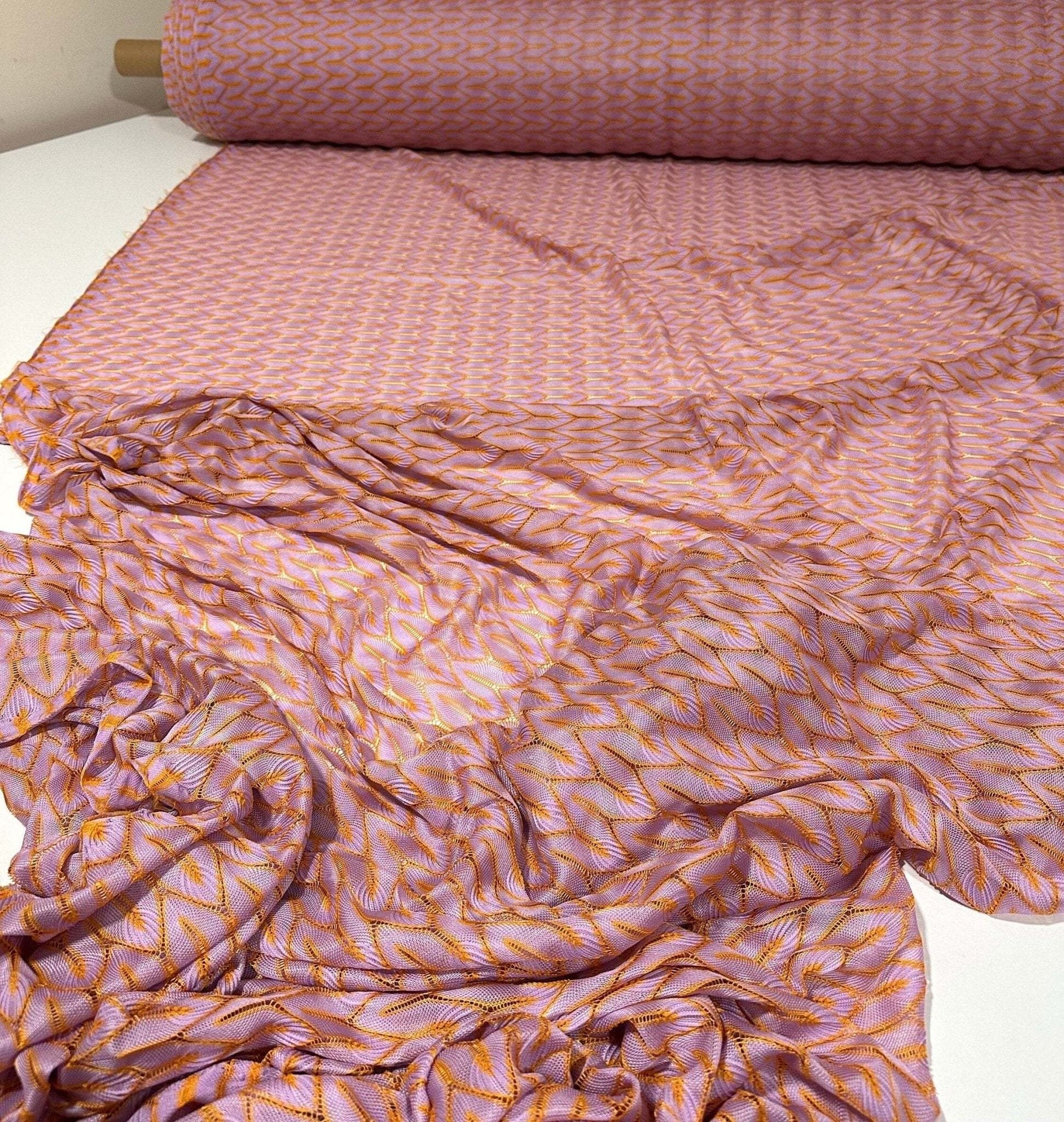 Roll of lilac and orange zigzag knit jersey mesh fabric spread out on white surface for crafting and fashion projects