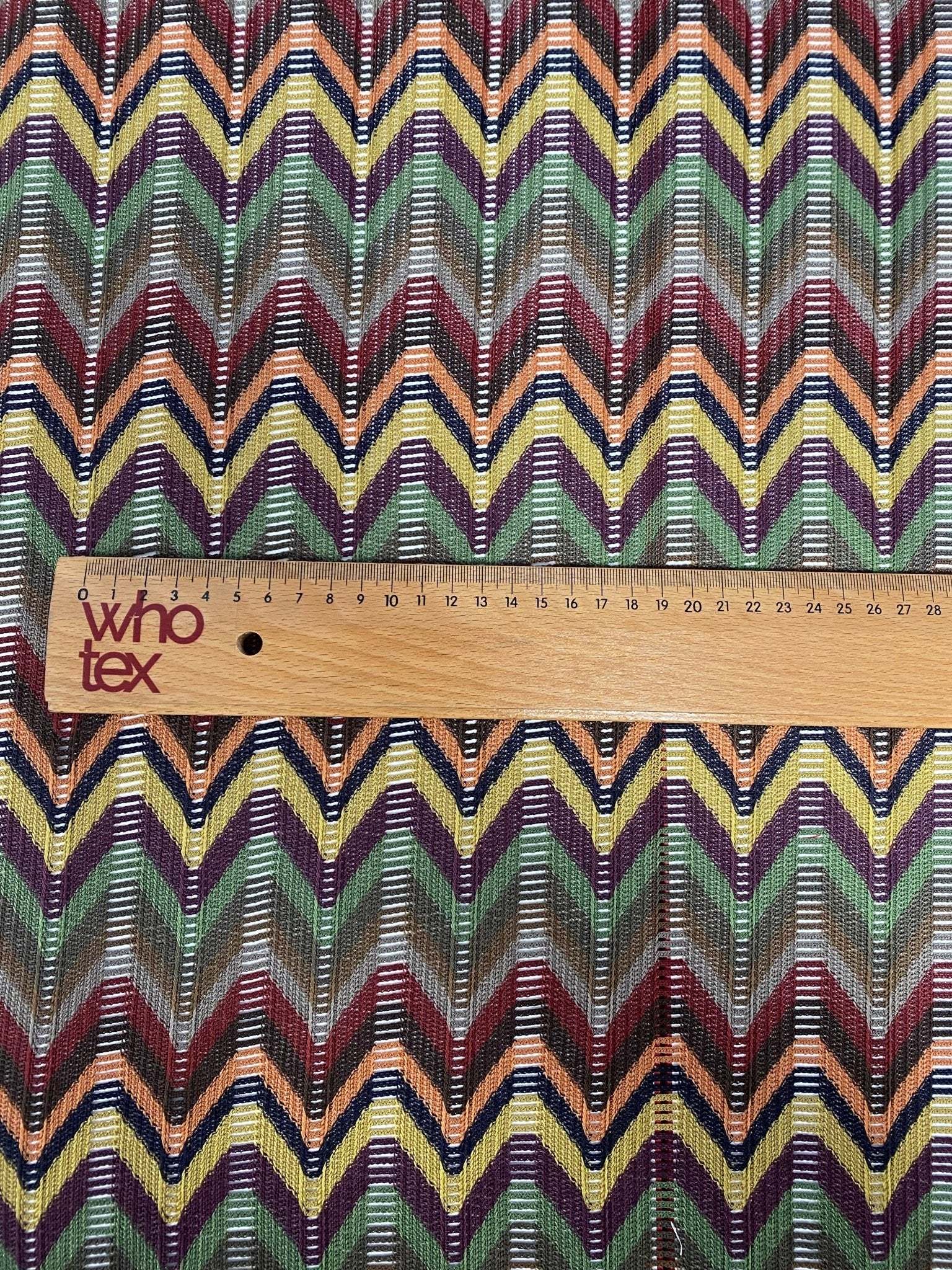 Multicolor Chevron Jacquard Fabric with zigzag pattern in green, yellow, red, and purple tones shown with wooden ruler for scale