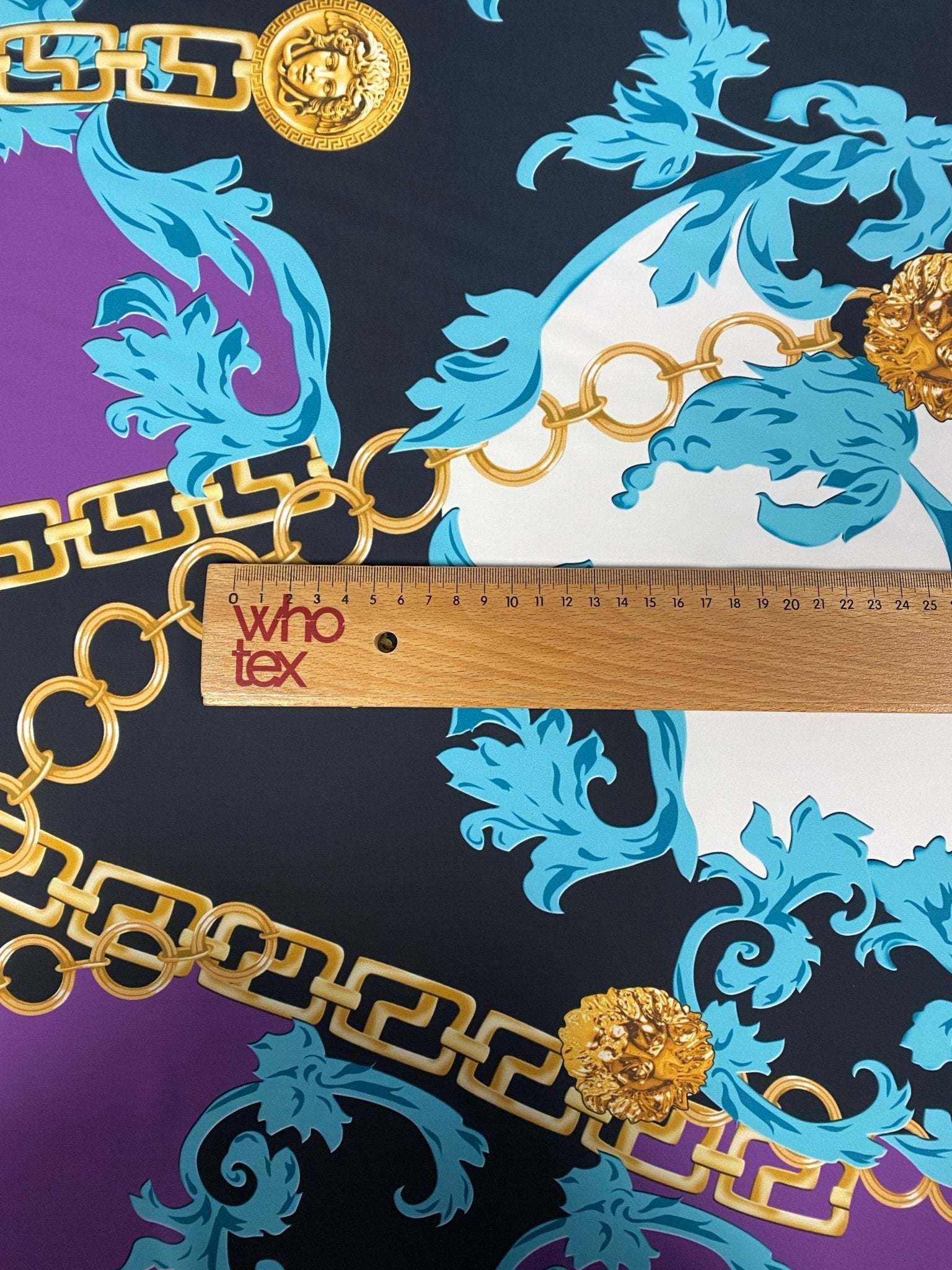 Swirl pattern crepe fabric with gold chain and blue floral designs on dark background next to wooden ruler