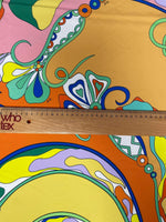 Close-up of vibrant psychedelic pattern fabric with bright orange, yellow, pink, green, and blue swirls and shapes, shown with a wooden ruler for scale.